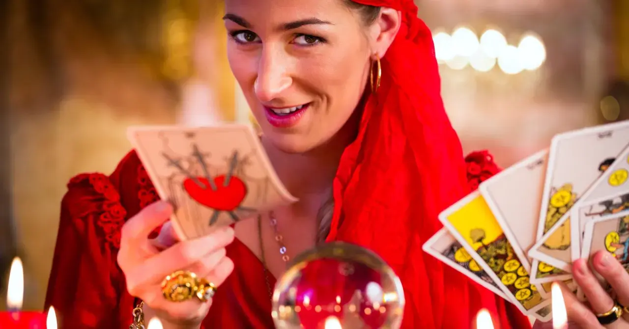 Kobieta w czerwieni trzyma karty tarota, zapowiadając baran tarot tygodniowy. Kryształowa kula i świece tworzą mistyczną atmosferę.