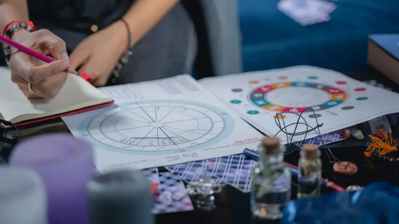 Osoba pisze w notatniku, obok wykresy astrologiczne, świece i talizmany. Analiza 12 dom astrologia.