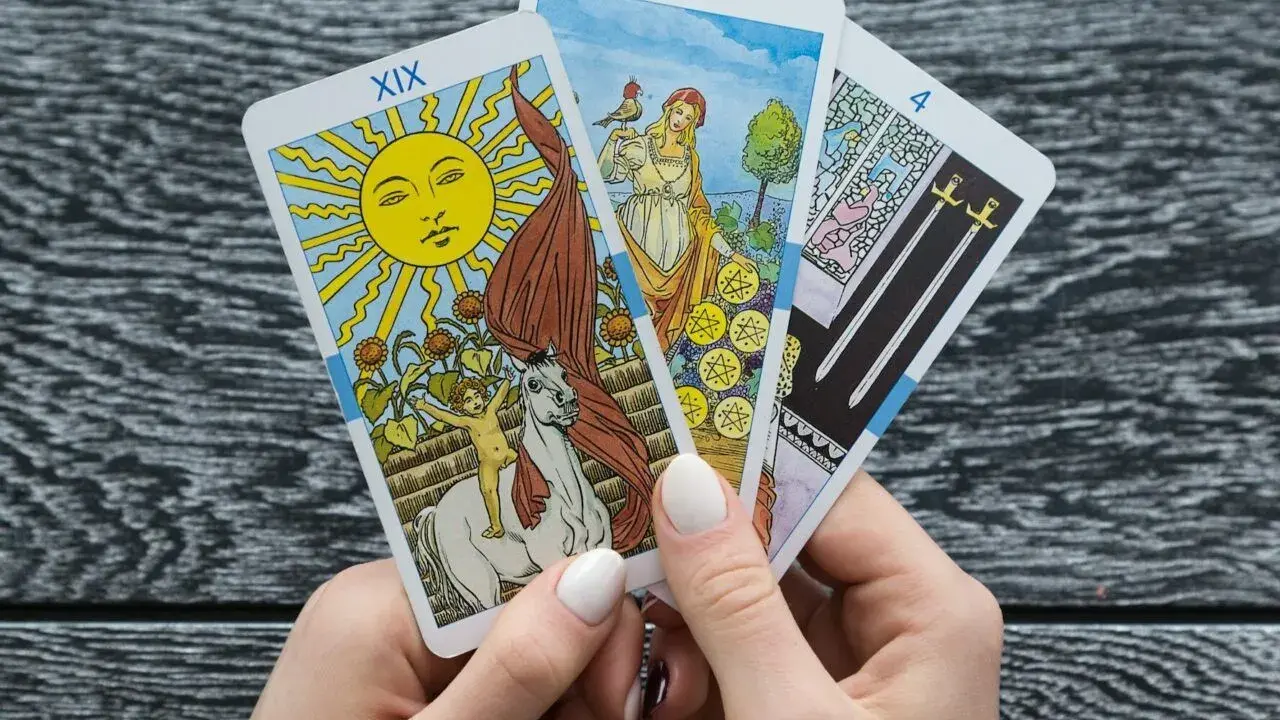 Ręce trzymają karty tarota: Słońce, Dziewiątka Monet i Czwórka Mieczy. To może być tarot miesięczny dla Raka.