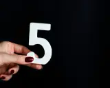 Dłoń z ciemnymi paznokciami trzyma białą cyfrę 5, symbolizującą numerologiczna 5 mężczyzna, na czarnym tle.