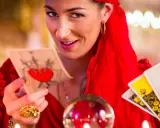 Kobieta w czerwieni trzyma karty tarota, zapowiadając baran tarot tygodniowy. Kryształowa kula i świece tworzą mistyczną atmosferę.