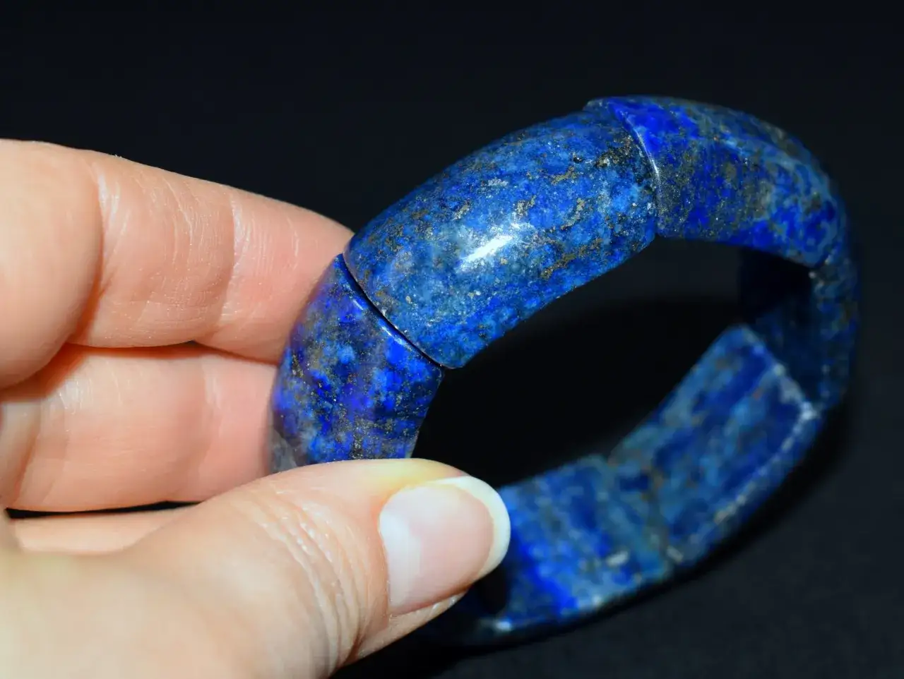 Dłoń trzyma bransoletę z lapis lazuli. Kamień ten, znany ze swoich właściwości, zachwyca głębokim niebieskim kolorem i złotymi inkluzjami.
